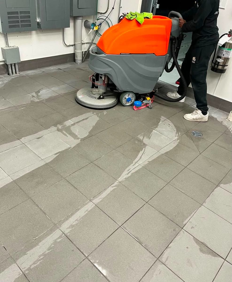Cleaner operating a ride-behind floor scrubber on a grey tiled floor